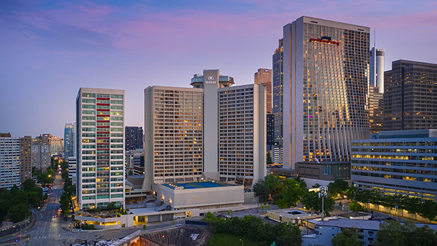 An aerial view of the Hilton Atlanta at dusk, showing the hotel's multi-tower complex surrounded by downtown Atlanta skyscrapers and tree-lined streets lit by warm evening light.