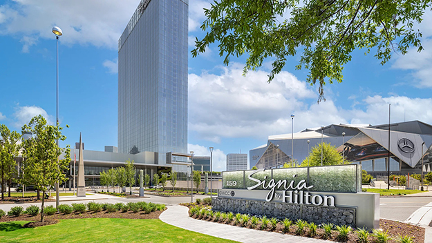 A ground-level view of the Signia by Hilton Atlanta entrance sign and landscaped walkway, with the hotel's glass tower rising behind it and Mercedes-Benz Stadium visible in the background.