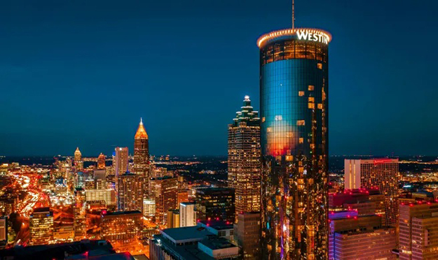 The cylindrical Westin Peachtree Plaza tower stands prominently above the glowing Atlanta skyline at night, with the Westin logo illuminated near the top and city lights stretching to the horizon.