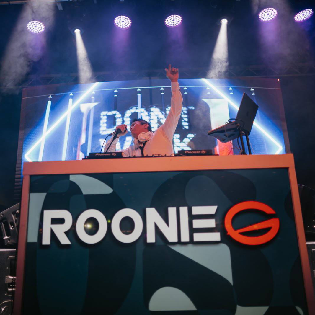 A man wearing a white shirt stands behind a DJ booth with his left hand raised into the air while holding a microphone with his right. The DJ facade has the logo for DJ Roonie G.