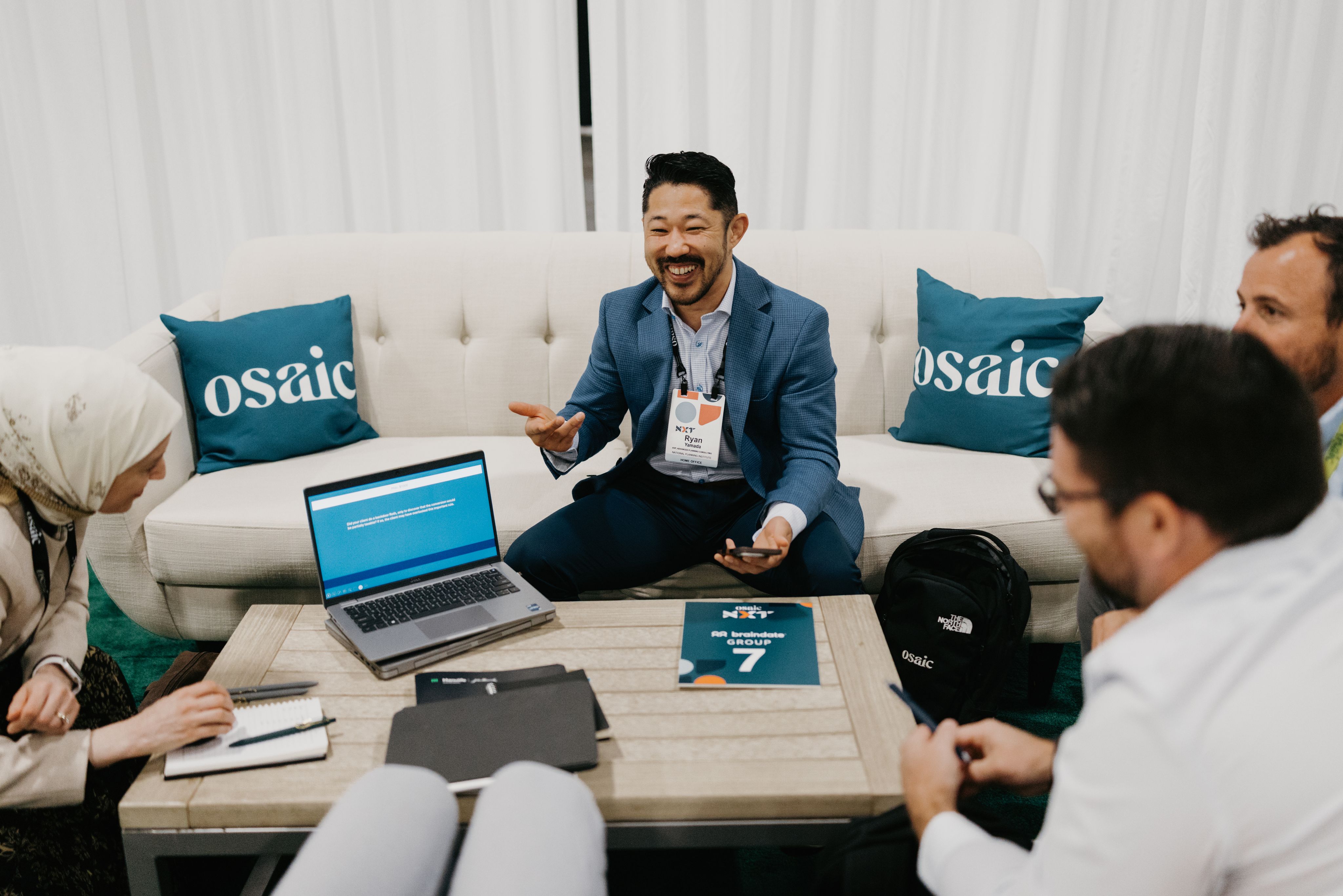 Image of Braindate Session. Group of 3 men sitting on a rounded couch in discussion. Teal pillows are sitting on the couch with an Osaic logo. A laptop sits on the table.