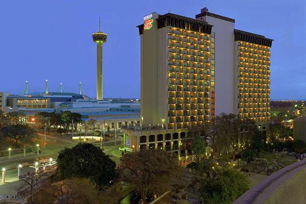 The Hilton Palacio del Rio building is lit up by lights on most of its 21 stories. Several trees are lined up in front of the hotel in the bottom foreground. In the background is the The Tower of the Americas observation tower.