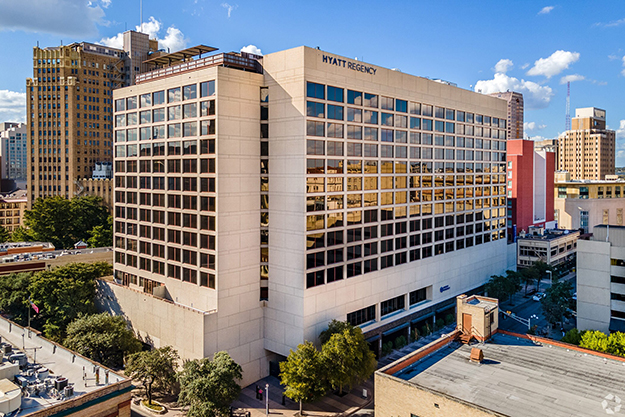 The Hyatt Regency Riverwalk hotel building is shown close up. One side shows a bright sunny reflection of buildings. Behind it, other buildings appear against a bright blue sky with a few clouds.