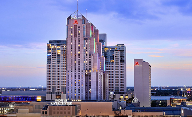 A handful of buildings tower above the skyline. Two have the bright neon red Marriott M logo near the top. A smaller building has a bright 'Shops at Rivercenter' sign affixed to its roof.