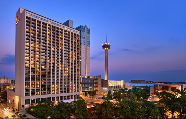 The Marriott Riverwalk stands at least 20 floors. The bright neon red Marriott M logo appears near the top. A little ways behind the building is the The Tower of the Americas observation tower.