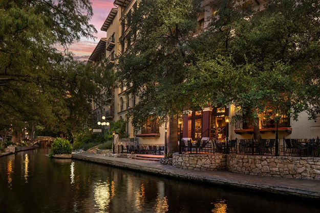 Big leafy tree branches stretch out from both sides of the San Antonio Riverwalk. On the right side, rows of windows of hotel rooms face the river, which stretches back into the distance and curves around the sidewalk.