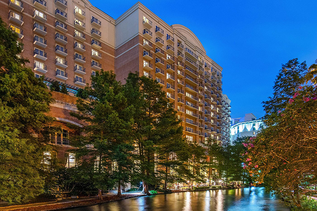The side of the building shows rows of balconies and windowed doorways leading into hotel rooms. Several trees cover the front of the building. In front of the sidewalk is a river reflecting the hotel lights.